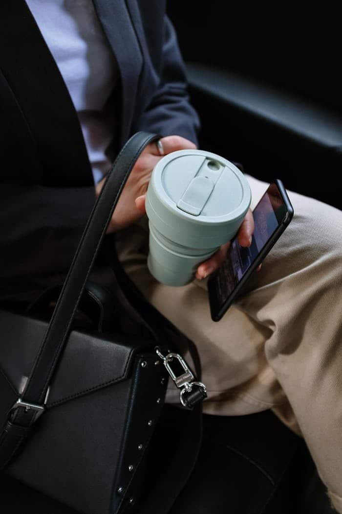 Persona con pantalones marrones sentada en asiento trasero de taxi de cuero negro con café en la mano y teléfono