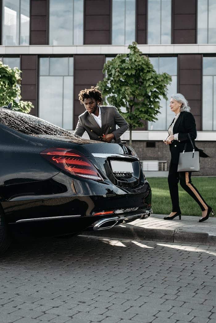 Un chofer con traje negro de pie junto a un coche negro abriendo la puerta a una clienta